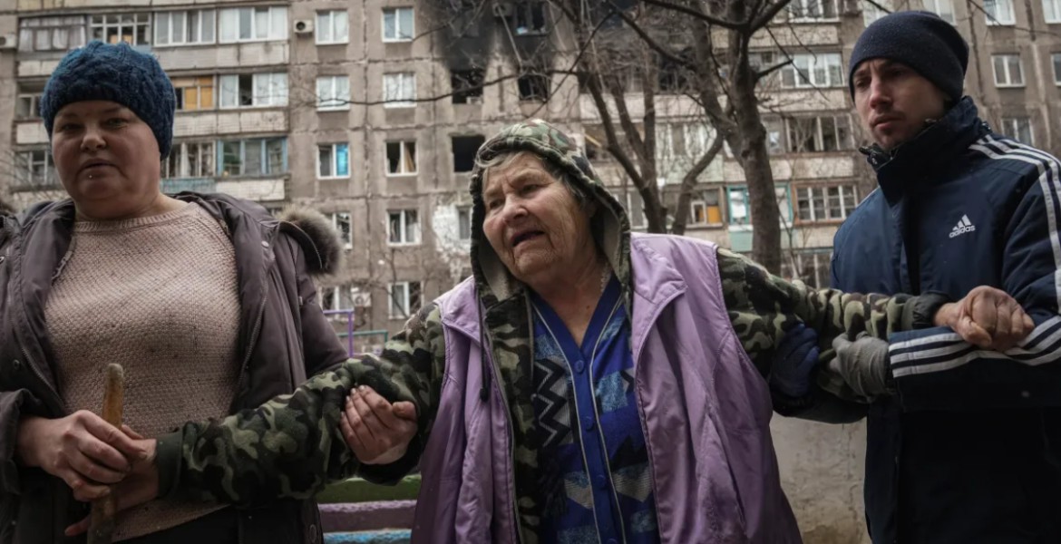 People help an elderly woman to walk in a street, in front of an apartment building hit by shelling in Mariupol, Ukraine. [Evgeniy Maloletka/AP Photo]
