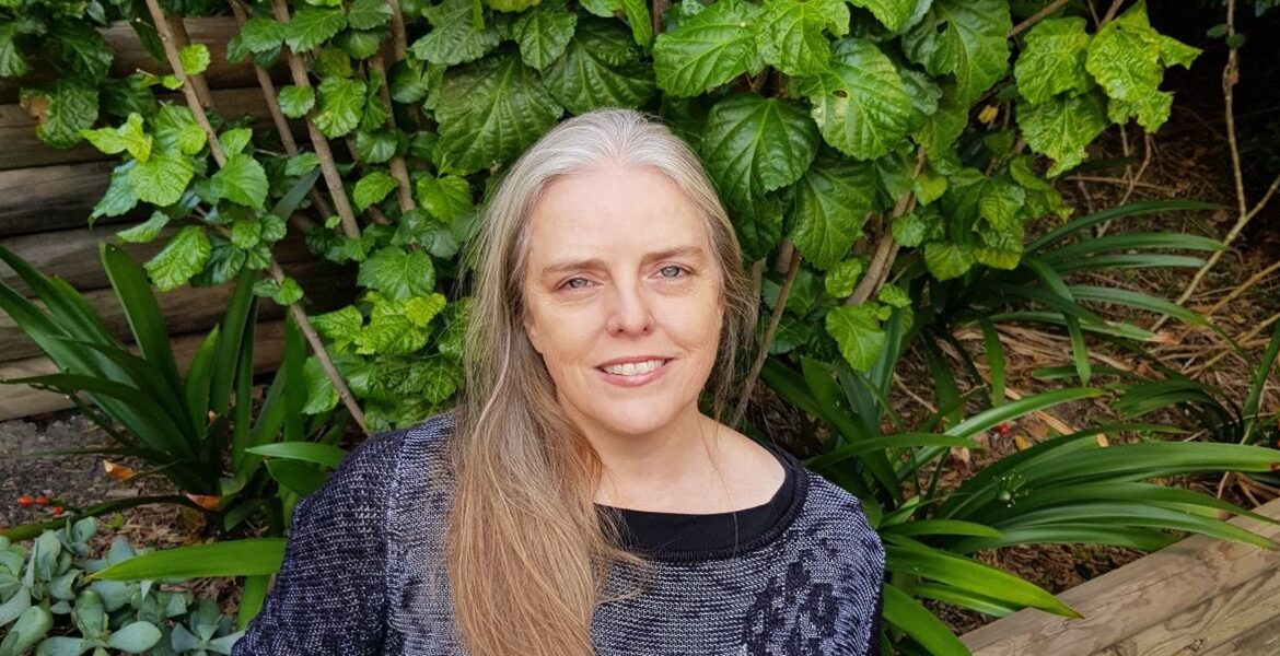 Author Sally Jane Smith. Image of a woman with long blond hair wearing a black top standing in front of a bush