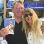 Author Peter Barber and his wife Alexandra. Image of a sandy color haired man wearing glasses and a black top holding up a book, the other arm around a woman with long blond hair wearing sunglasses and a while top