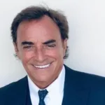 Actor Thaao Penghlis Cookbook Author Image by Thaao Penghlis. Image of a siling, tanned man with dark hair in a black suit, white short, and tie.