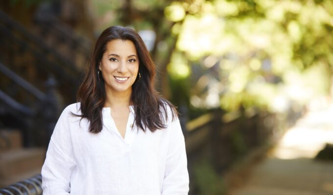 Author Yvette Manessis Corporon. Image by Connie Fernandez. Image of a woman with long brown hair wearing a white bouse standing near trees