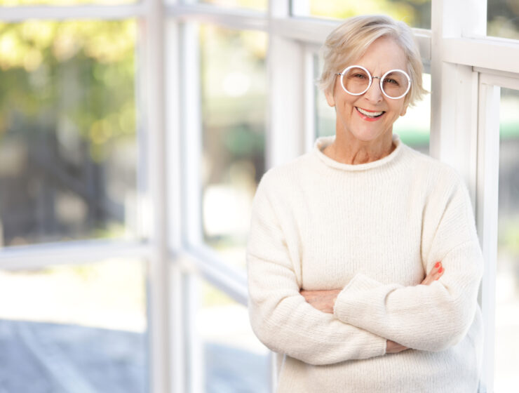 Photo of author Australian Author Shelley Dark. IMAGE BY BRISBANE HEADSHOTS. Image of a smiling woman with short blond hair, standing in front of a window and wearing large round, white glasses and a white sweater
