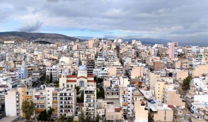 Aerial view of Athens showing a densely populated city with a mix of modern and traditional architecture, including a prominent church with a blue dome, under a cloudy sky.