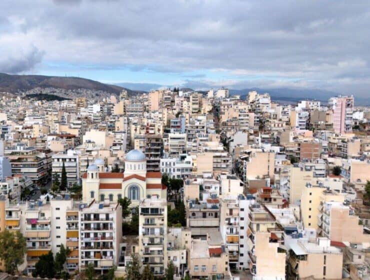 Aerial view of Athens showing a densely populated city with a mix of modern and traditional architecture, including a prominent church with a blue dome, under a cloudy sky.
