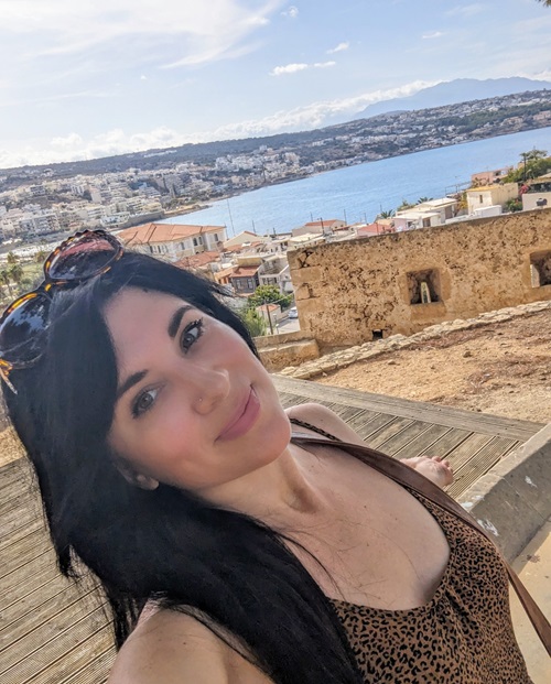 Chef Krystina Kalapothakos of Travel Mazi Mou. IMAGE: ©2025 Travel Mazi Mou/Krystina Kalapothakos. Image of a smiling woman with long black hair posing from a high elevation in Greece overlooking the sea