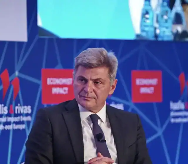 Thoughtful Greek politician during conference event, wearing formal suit, with a blue futuristic background displaying Greek and global themes.